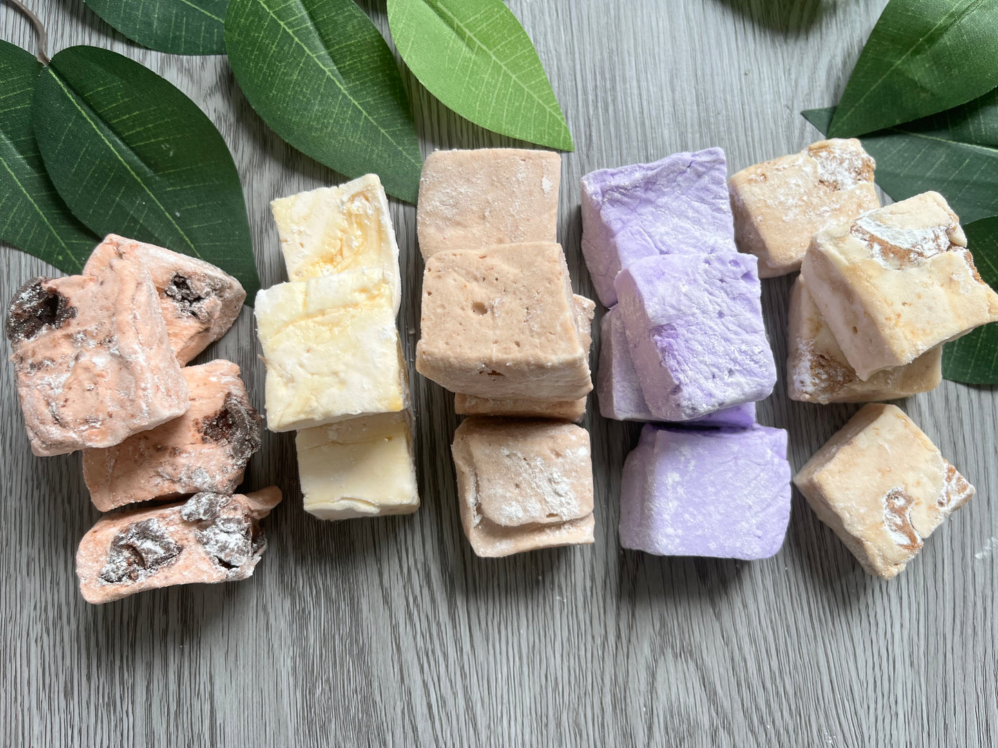 Assorted gourmet marshmallows in multiple flavors, cut into soft cubes and arranged on a wood board with green leaves — fluffy, colorful, and handmade with halal ingredients.