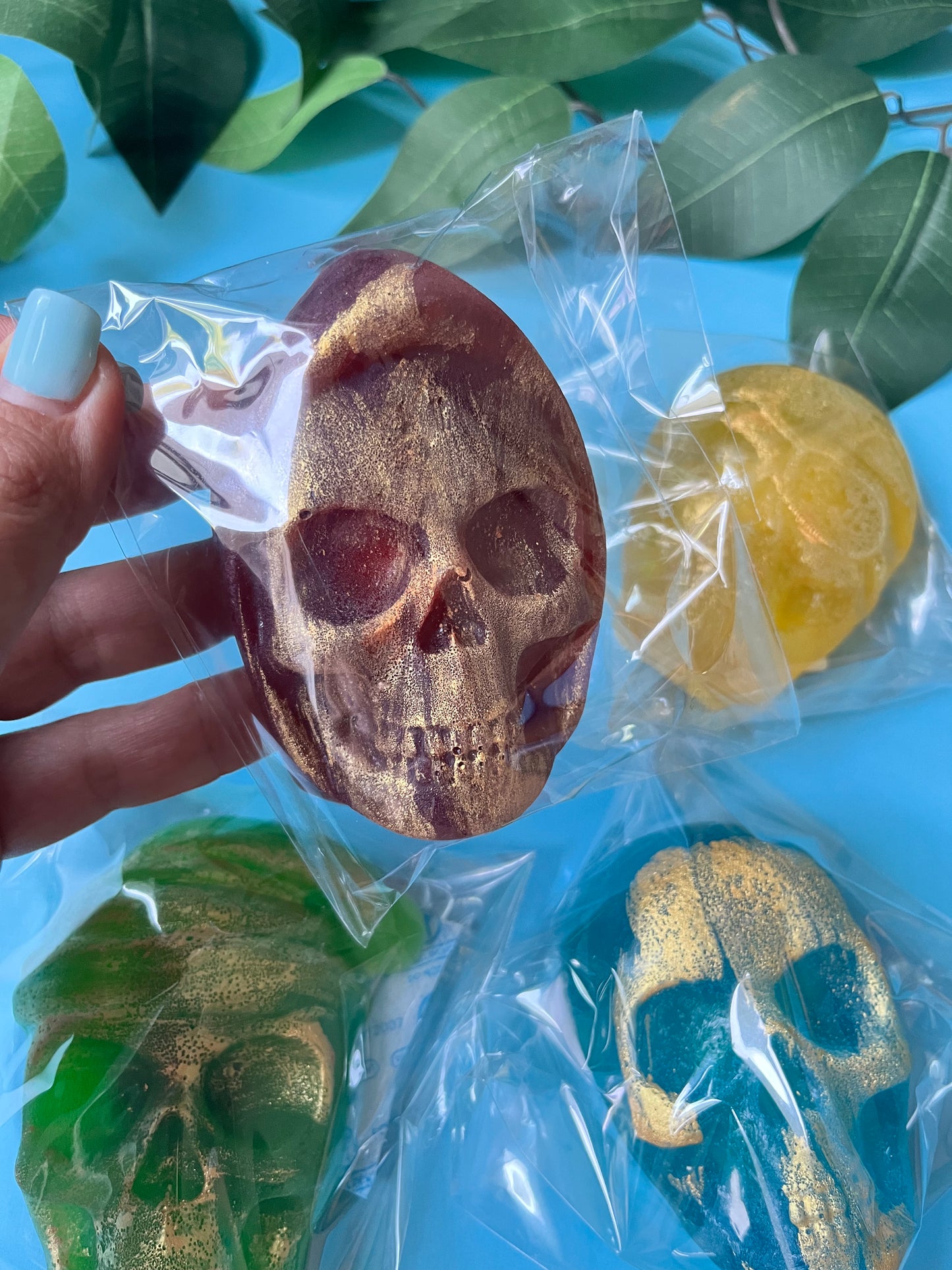 Skull-shaped soap with gold glitter held in front of other similar soaps on a blue background with green leaves.