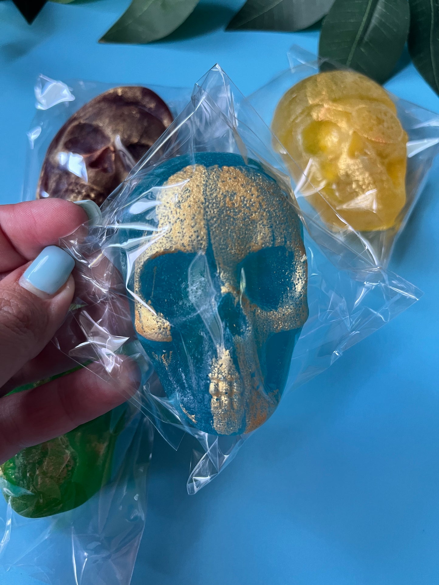 Hand holding a blue skull-shaped Crystal CAndy with gold detailing, surrounded by other bath bombs on a blue background.