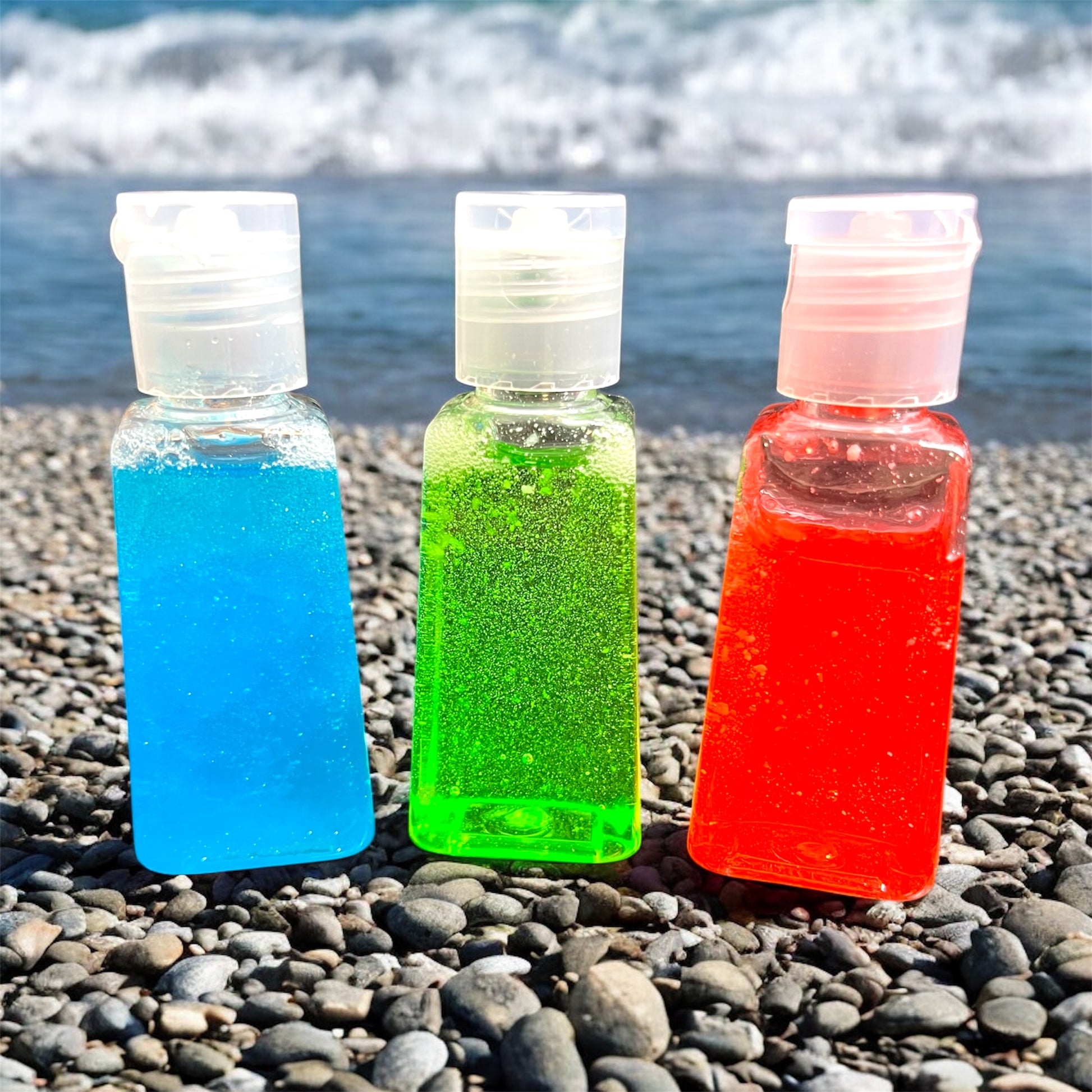 Three small bottles of blue, green, and red liquid on a pebbly beach.
