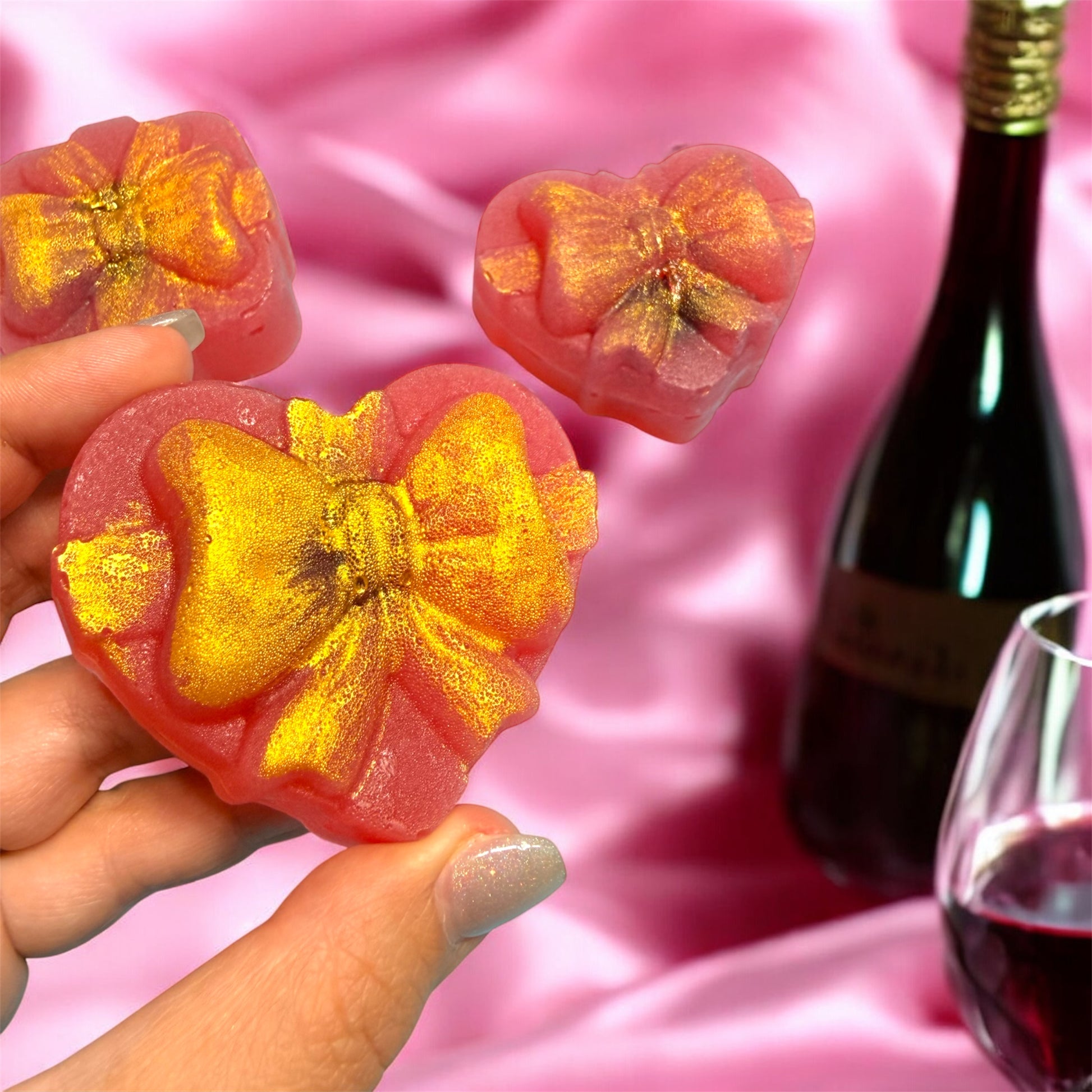Hand holding a pink flower-shaped dessert with gold glitter against a pink background with a bottle and glass of red wine.