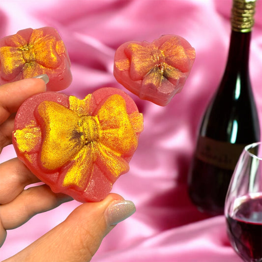 Hand holding a pink flower-shaped dessert with gold glitter against a pink background with a bottle and glass of red wine.