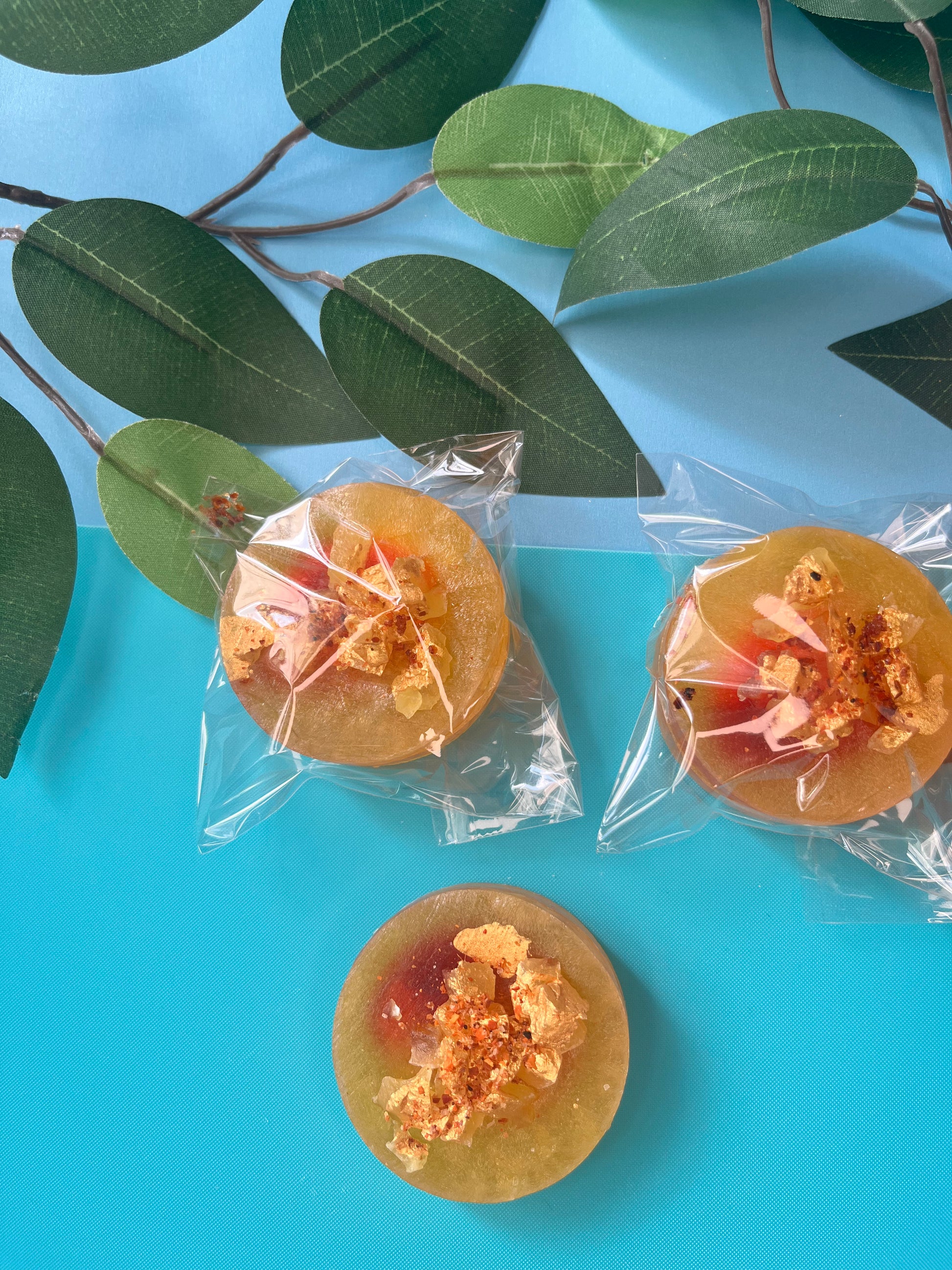 Top view of round mango crystal candies wrapped in clear plastic, each topped with golden crystal shards and a sprinkle of tajín, laid out on a light blue surface with leafy green props.