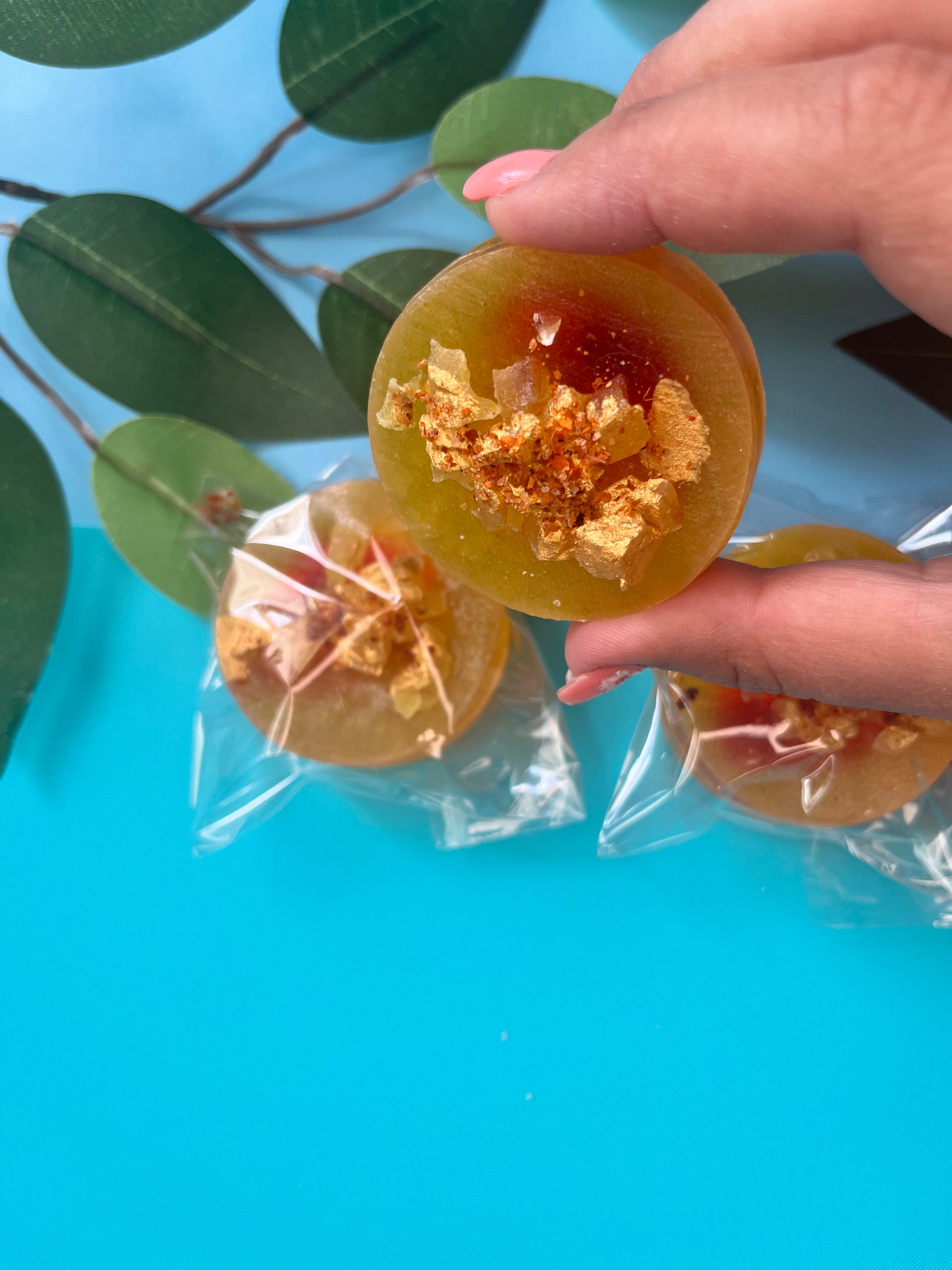 Hand holding a split mango crystal candy filled with red chamoy sauce, topped with golden mango candy shards and tajín, with more wrapped candies and green leaves in the background.
