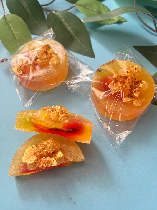 Close-up of an unwrapped mango crystal candy, showing glossy golden-orange shell, visible chamoy center, crunchy shards on top, and hand-painted gold detailing.