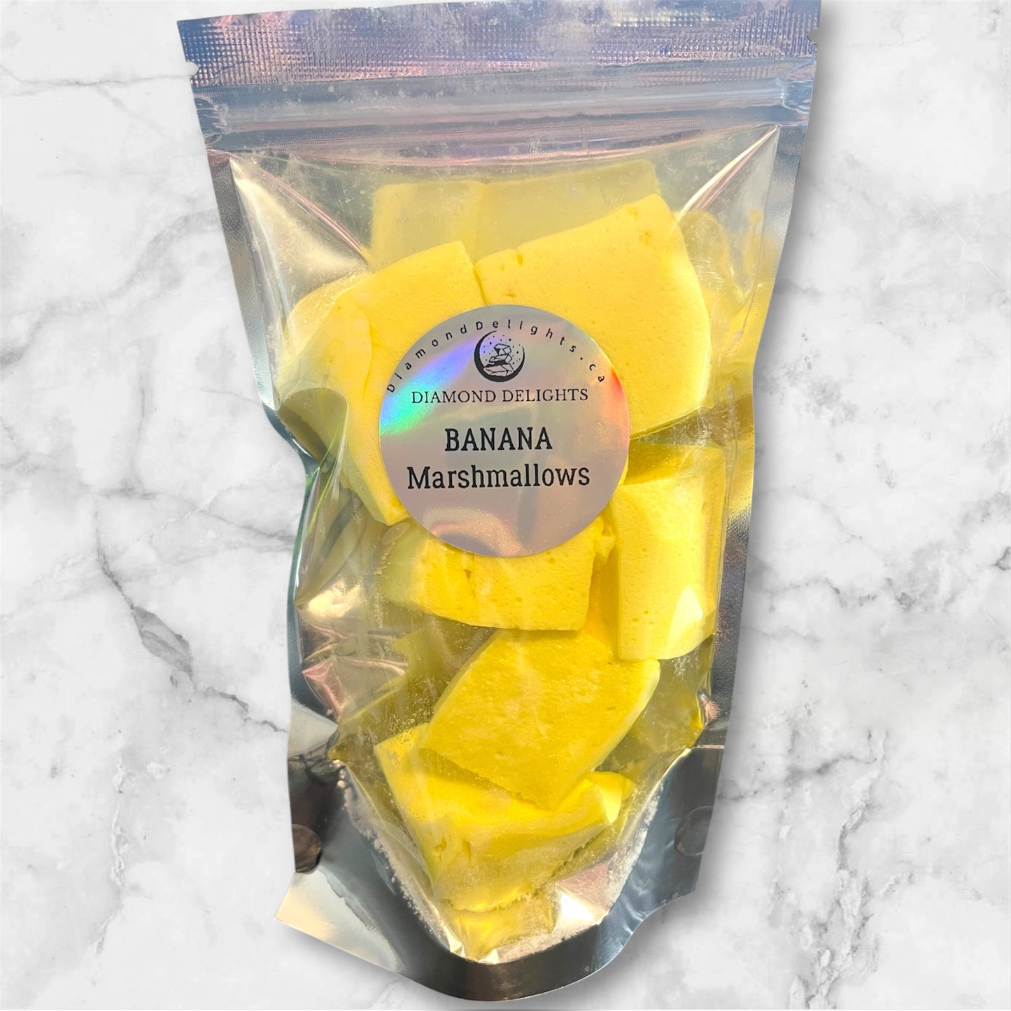 Bag of bright yellow banana marshmallows in a clear stand-up pouch with holographic Diamond Delights label, set against a marble background.