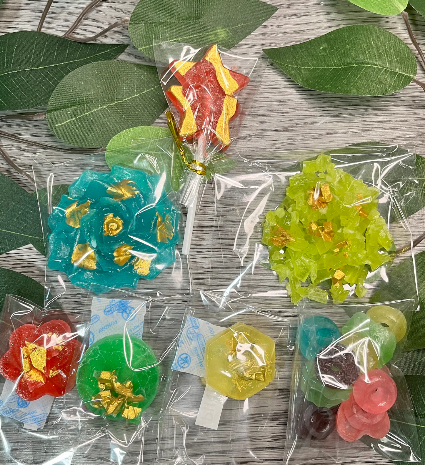 Assorted handmade crystal candies from Diamond Delights’ “EVERYTHING” Box, including a flower, lollipop, cluster, and Gemmies displayed on green leaves.