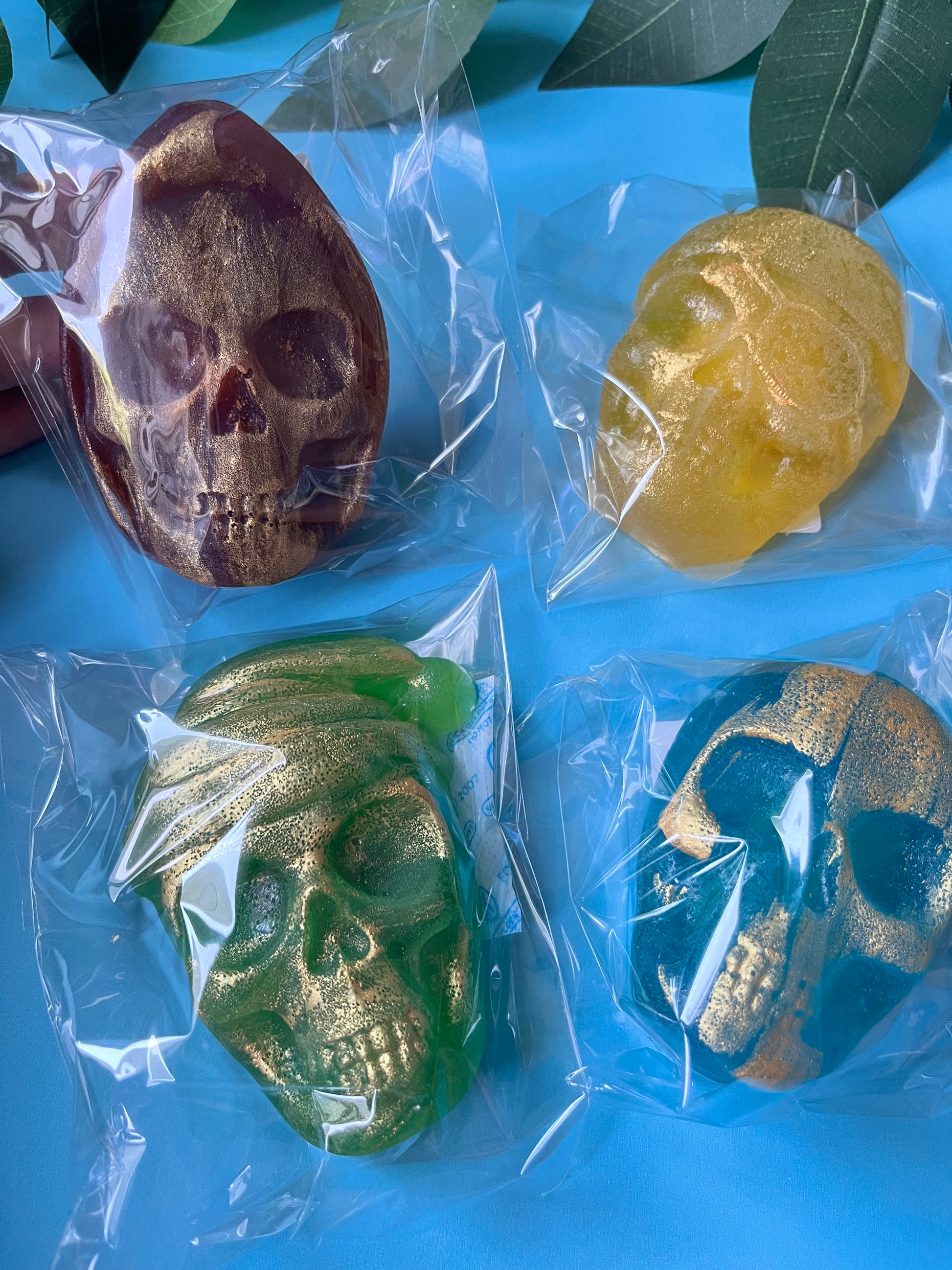 Four skull-shaped Crystal Candies in different colors (brown, yellow, green, blue) on a blue surface with leaves.