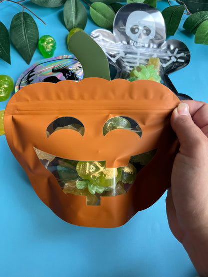 Hand holding a pumpkin-shaped candy bag filled with green candies against a blue background with skeleton and ghost decorations.