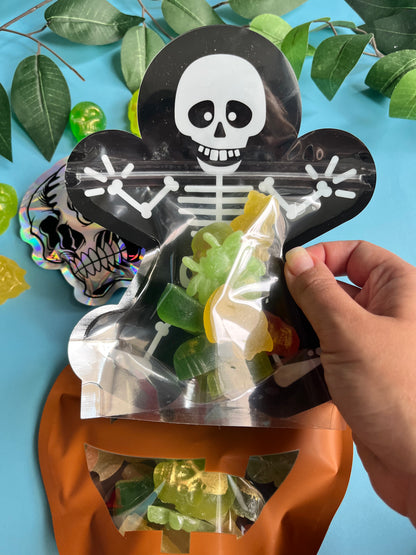 Hand holding a transparent bag of gummy candies with a skull and crossbones design on a blue background with green leaves.