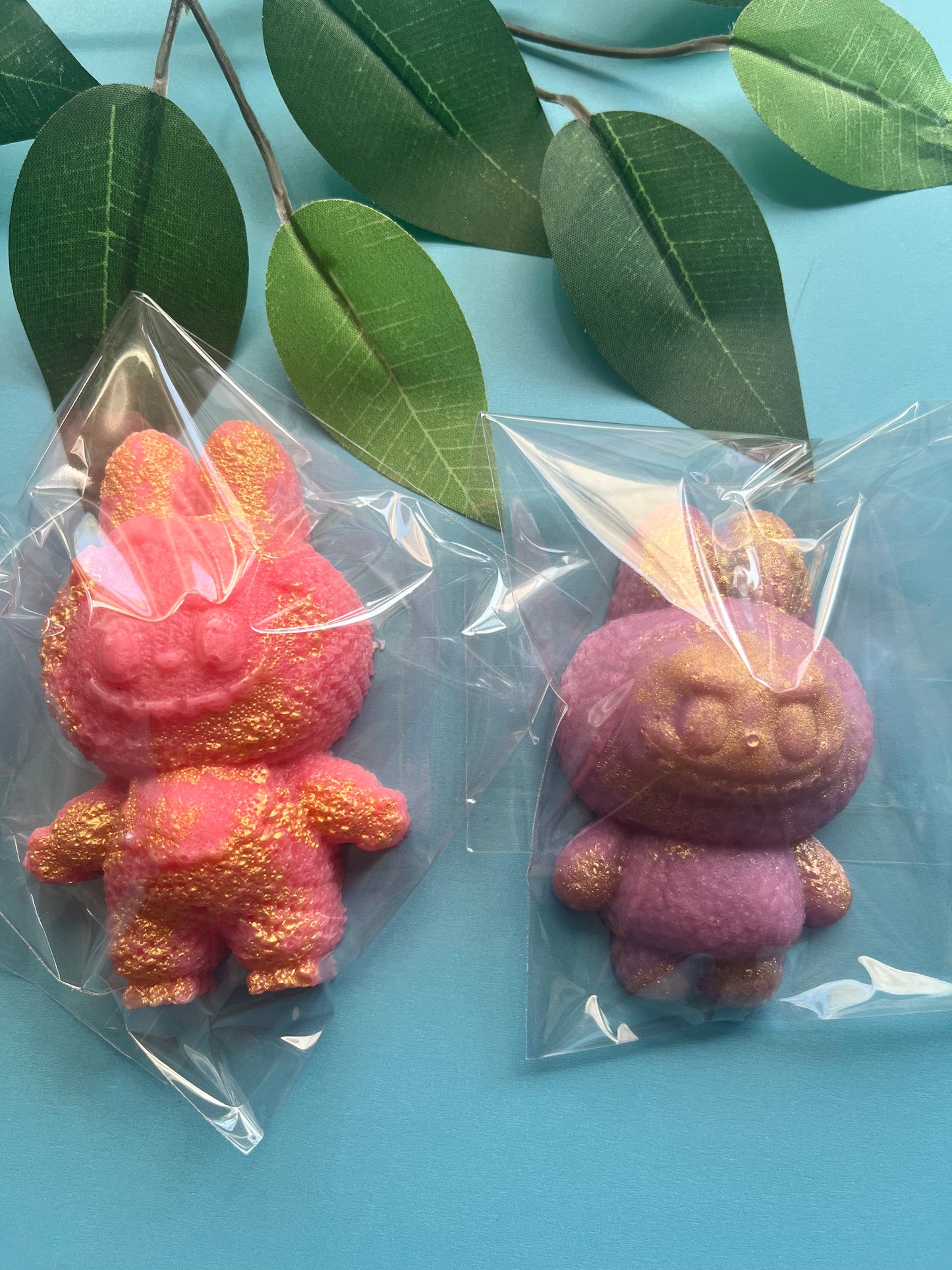 Two crystal candy monsters, one pink and one purple, in clear plastic bags on a blue background with green leaves.
