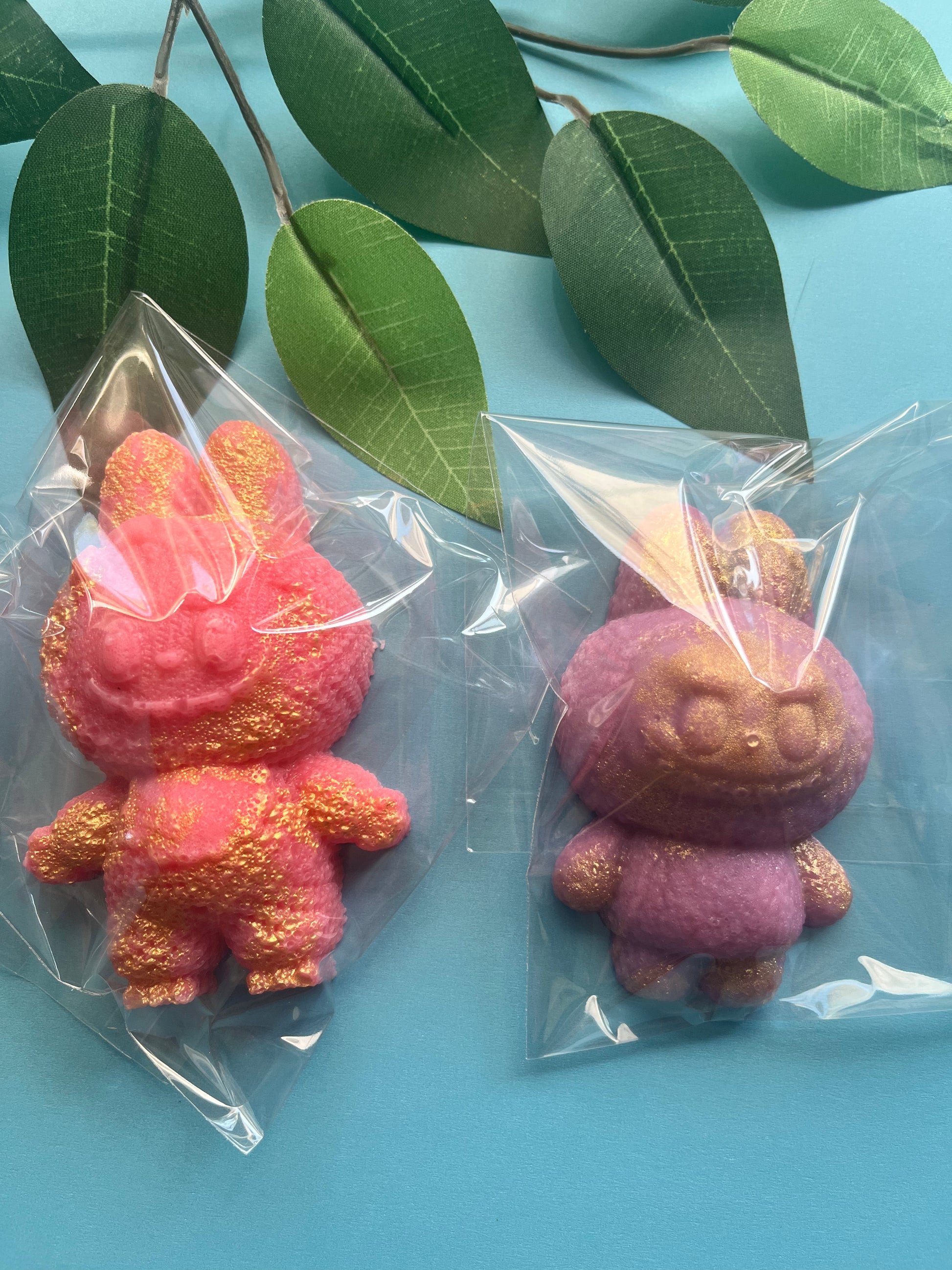 Two crystal candy monsters, one pink and one purple, in clear plastic bags on a blue background with green leaves.